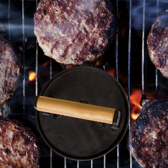 Five burger patties cook on a grill, with the Jim Beam Cast Iron Burger Press—featuring a wooden handle—on one patty. Flames and hot coals glow below, creating an ideal scene for any grilling enthusiast or cast iron cookware fan.