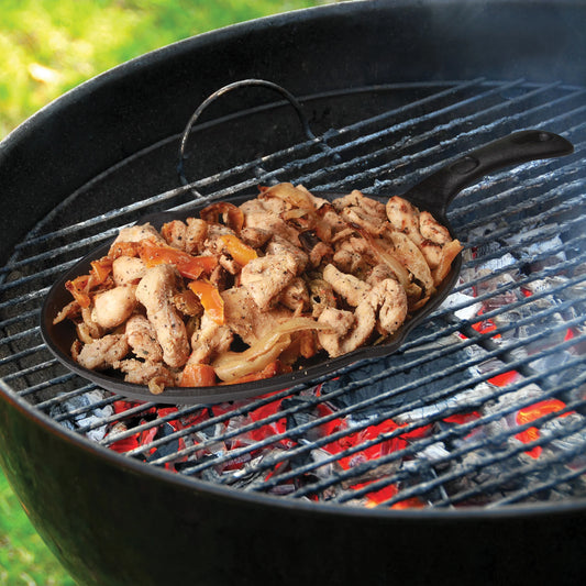 The Jim Beam Cast Iron Skillet Set from Jim Beam is shown filled with chicken and vegetables, cooking over hot coals. Smoke rises as it cooks, demonstrating this pre-seasoned cookware’s functionality for outdoor grilling.