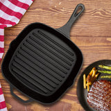 The Jim Beam Cast Iron Square Grill Pan, known for excellent heat retention, rests on a wooden surface beside a red and white checkered cloth and a plate of steak, asparagus, and roasted potatoes.