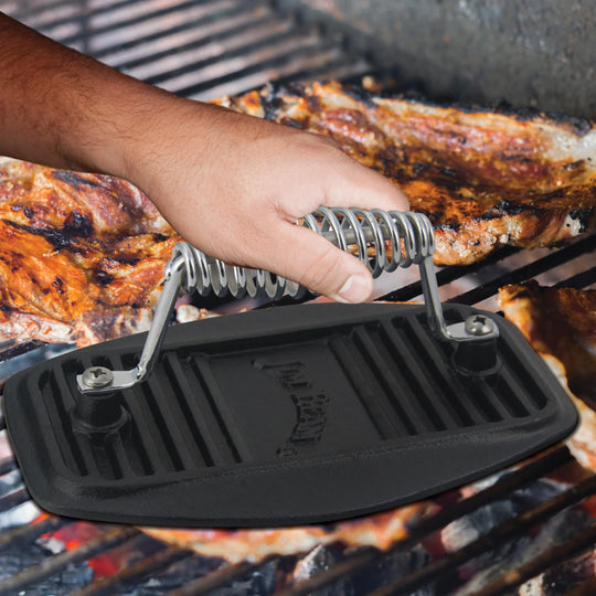 The Jim Beam Cast Iron Meat Press, shown with a metal coil handle and ribbed underside, is an essential grilling tool for pressing food evenly while cooking.