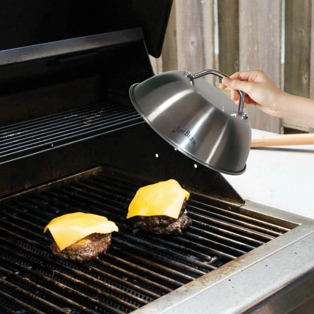 A hand lifts the Jim Beam Stainless Steel 9 Burger Dome by Jim Beam to reveal perfectly melted cheese on two burgers grilling, with an open grill and wooden fence in the background for the ultimate cookout.