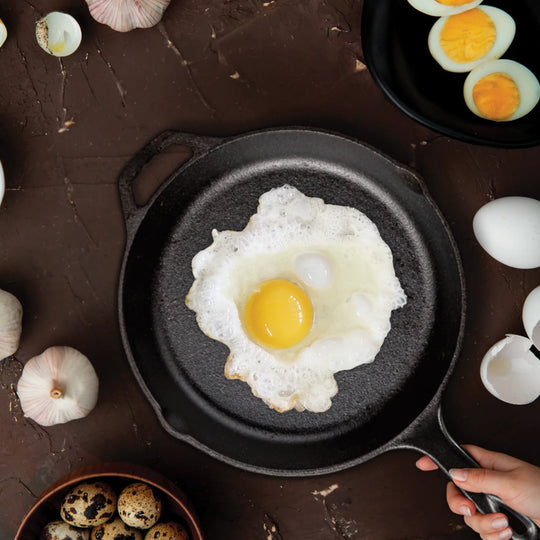 A fried egg sizzles in the Jim Beam Cast Iron 10.25 Skillet, surrounded by garlic, a bowl of quail eggs, and eggshells. A hand grips the handle—highlighting this Jim Beam essential for every kitchen collection.