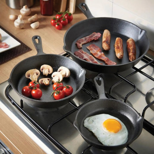 Three Jim Beam Cast Iron skillets from the Jim Beam 3 Piece Skillet Set hold cherry tomatoes with mushrooms, bacon and sausages, and a sunny-side up egg on the stove. Fresh vegetables rest on the nearby countertop.