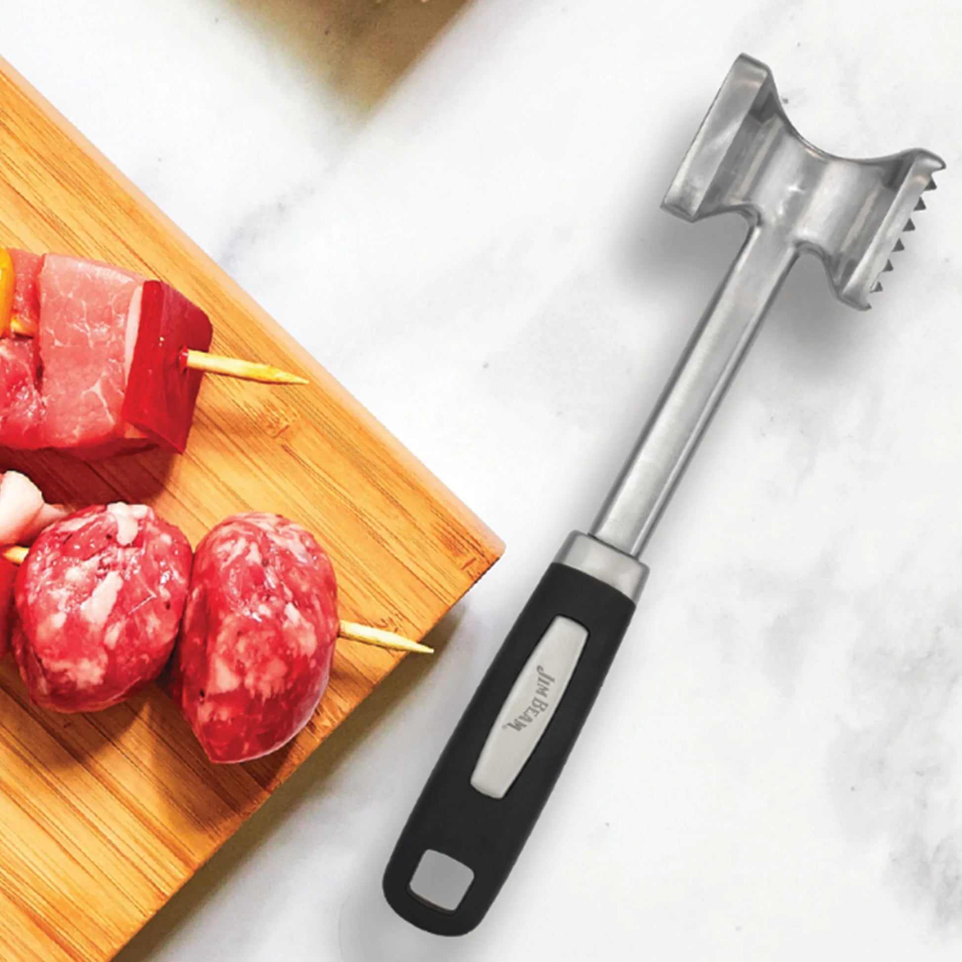 The Jim Beam Dual Side Meat Tenderizer by Jim Beam, featuring a black handle, rests on a white surface next to a wooden cutting board with raw meat skewers.