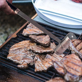 A close-up of grilled steaks on a barbecue, with a hand holding a knife and tenderizer over them, and a white plate with raw meat and the Jim Beam Salt and Pepper Shaker by Jim Beam in the background.
