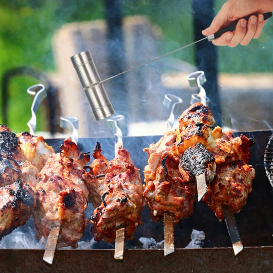 Several skewers of grilled meat cook over an open flame on a barbecue. A hand seasons the meat with the Jim Beam Salt and Pepper Shaker by Jim Beam, while smoke rises and the outdoor background remains blurred.