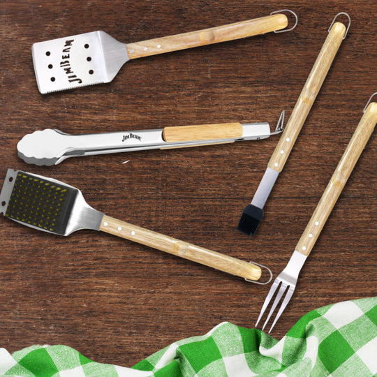 The Jim Beam 5-Piece Grill Tool Set includes stainless steel tools with wooden handles—spatula, tongs, basting brush, fork, and a bonus tool—displayed on dark wood beside a green-and-white checkered cloth. Grill like a pro with Jim Beam!.