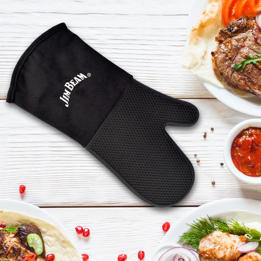 A Jim Beam Silicone Grilling Mitten by Jim Beam rests on a white wooden table, surrounded by grilled steak, sauce, wraps, herbs, and scattered pomegranate seeds.