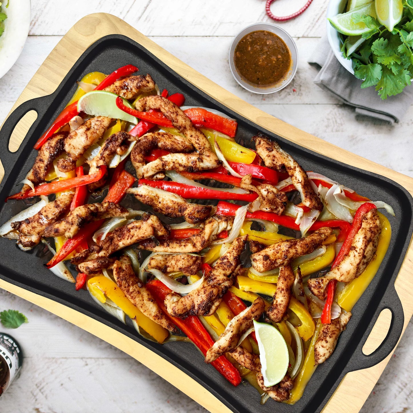 Grilled chicken fajitas sizzle in the Jim Beam Cast Iron Grill Pan Set by Jim Beam, with bell peppers, onions, and lime wedges. Sauce, cilantro, and extra lime sit nearby on a white table.