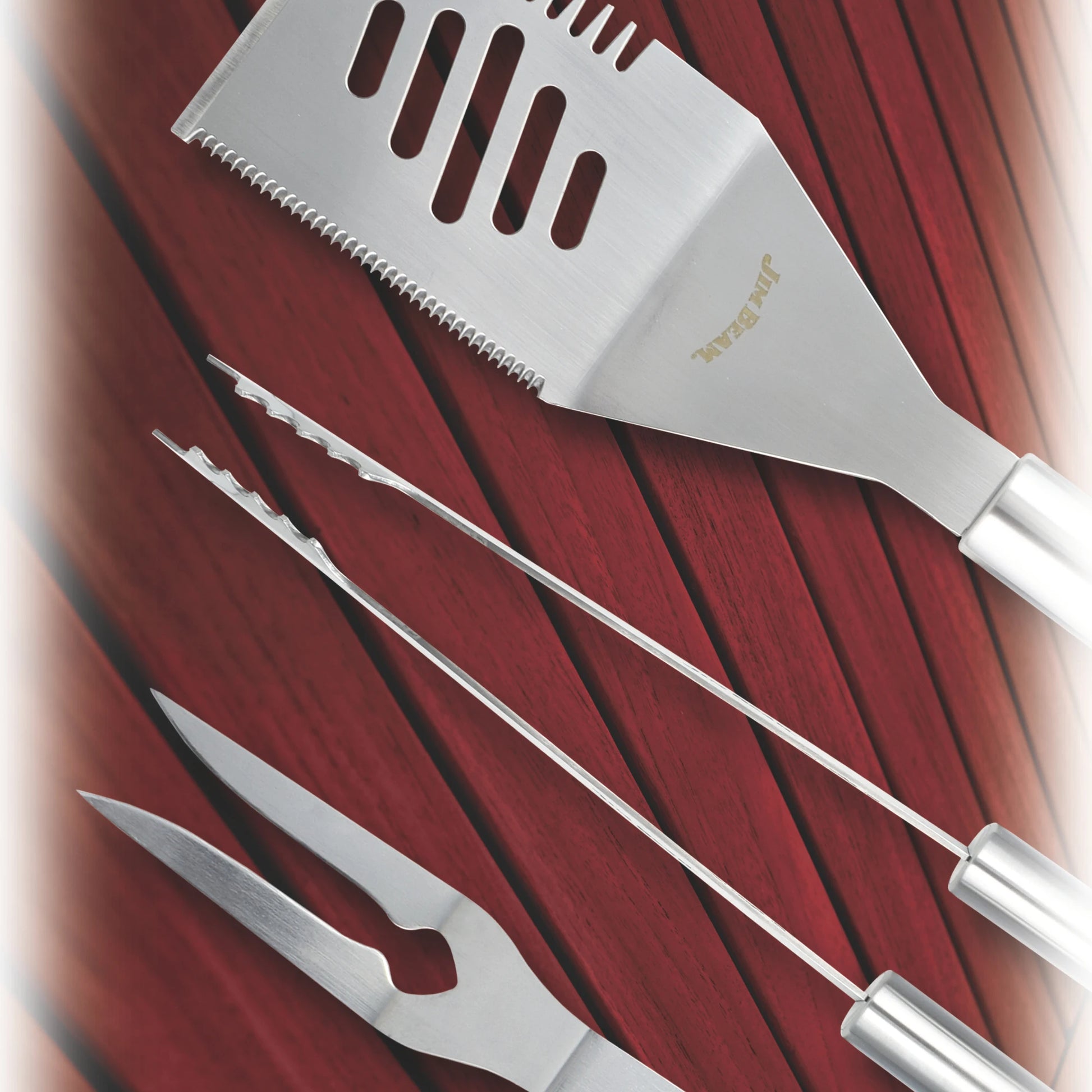 The Jim Beam 3 Piece BBQ Tool Set features stainless steel spatula, tongs, and Grilling BBQ Fork—each with pointed tips or serrated edges—arranged diagonally on a red wooden surface. The set is by Jim Beam.