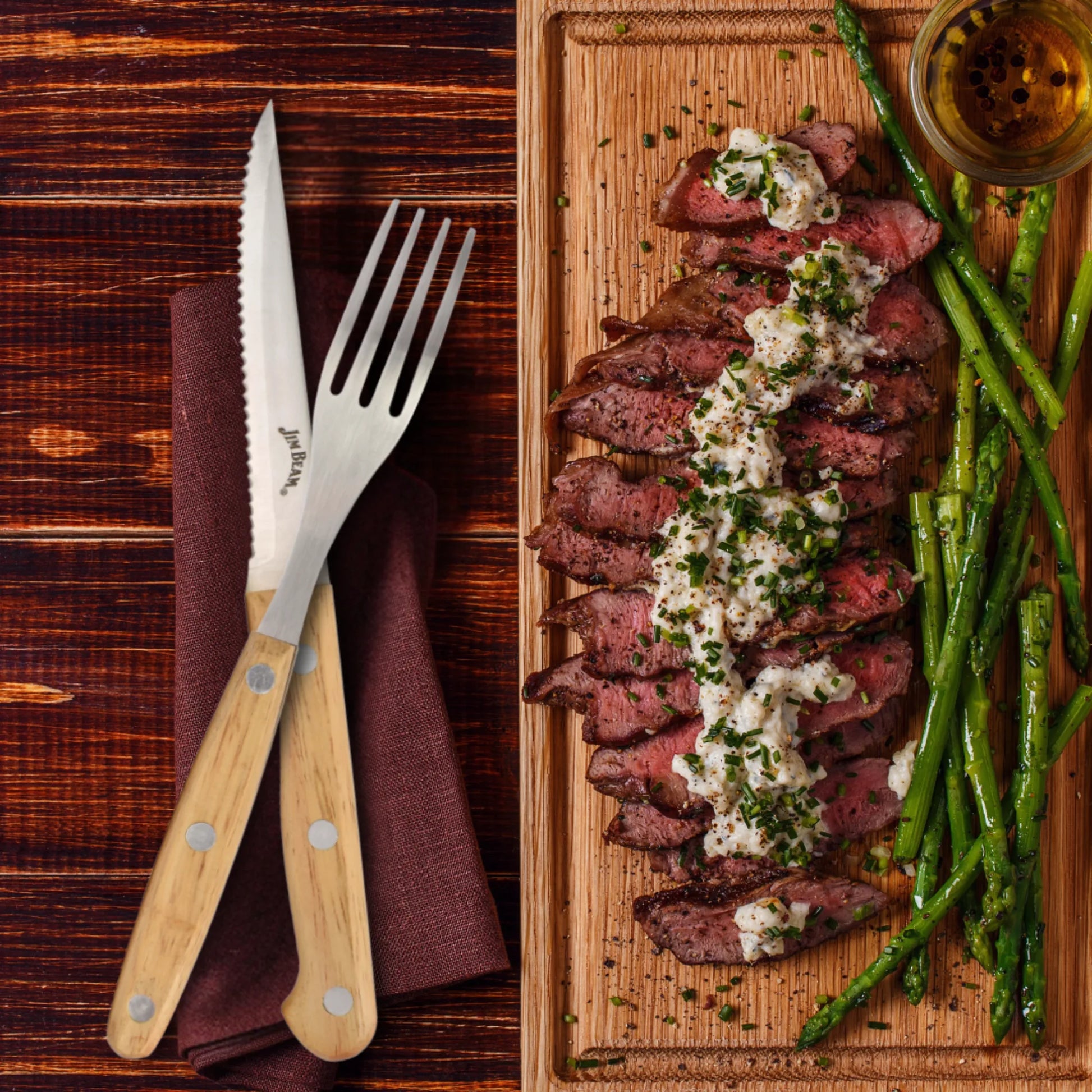 A wooden board holds sliced steak with creamy sauce and herbs, served with grilled asparagus. A Jim Beam Steak Knife and Fork from the Jim Beam 8 Piece Set rest on a brown napkin beside the board atop a dark wood table.