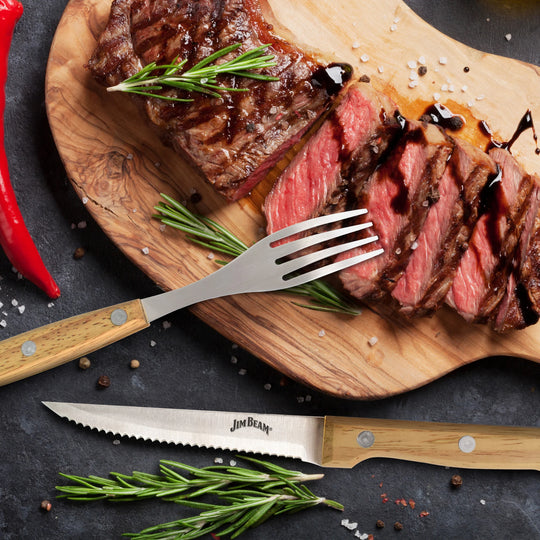 A sliced grilled steak, garnished with rosemary and sauce, rests on a wooden board beside red chili peppers, salt, and a fork and serrated steak knife from the Jim Beam 8 Piece Steak Knife and Fork Set by Jim Beam.