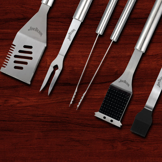 The Jim Beam 5-Piece BBQ Tool Set, featuring a spatula, fork, tongs, grill brush, and basting brush with engraved Jim Beam logos on each handle, is arranged side by side on a dark wood surface.