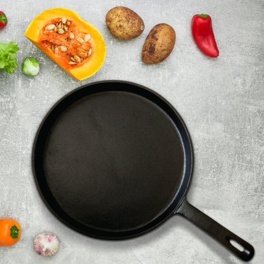 The Jim Beam Cast Iron 10.5 Griddle rests on a gray surface, surrounded by fresh vegetables like pumpkin, potatoes, chili pepper, tomato, lettuce, and a small onion.