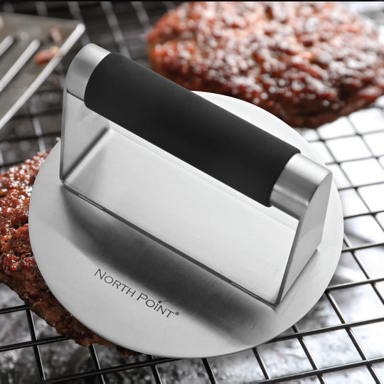 The North Point 3 Piece Burger Kit includes a stainless steel burger press with a black handle, shown on a wire rack beside cooked patties—a perfect kit for burger enthusiasts.
