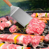 A close-up of raw meat and vegetable skewers cooking outdoors as food is expertly flipped with a spatula from the North Point 8 Piece Griddle Set, with smoke rising in the bright summer scene.