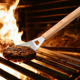 A spatula from the North Point 3 Piece BBQ Tool Set lifts a grilled beef patty from a hot grill with flames in the background, capturing the outdoor barbecue experience.