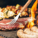 A North Point stainless steel BBQ tool from the North Point 3 Piece BBQ Tool Set presses a steak on the grill, while sausages and skewered vegetables cook alongside over an open flame.