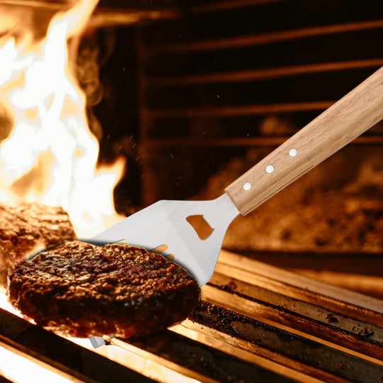 A hamburger patty is lifted from a grill with a stainless steel tool from the North Point 5 Piece BBQ Tool Set, featuring a wooden handle, as flames and another patty sizzle in the background.