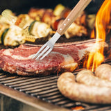 A close-up shows a steak held with a fork from the North Point 5 Piece BBQ Tool Set on the grill, with flames nearby. Sausages and skewers of grilled vegetables also cook over the heat. Brand: North Point.