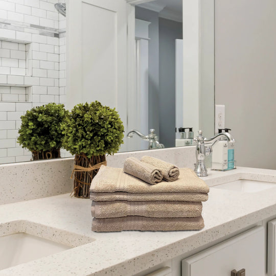 The Textilia Set of 6 Bath Towel Set – Beige from Textilia is neatly folded on a white countertop beside green plants, a soap dispenser, and a chrome faucet, with a mirror and tiled shower in the background.