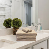The Textilia Set of 6 Bath Towel Set – Beige from Textilia is neatly folded on a white countertop beside green plants, a soap dispenser, and a chrome faucet, with a mirror and tiled shower in the background.