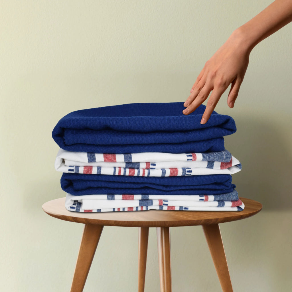 A hand reaches for the Textilia 4 Piece Kitchen Towel Set by Textilia, featuring 100% cotton patterned white and solid blue quick-dry towels stacked on a wooden stool against a light green wall.