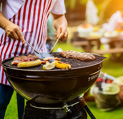 A person in a red-striped apron grills various meats and vegetables outdoors, surrounded by a vibrant green lawn and picnic setup.